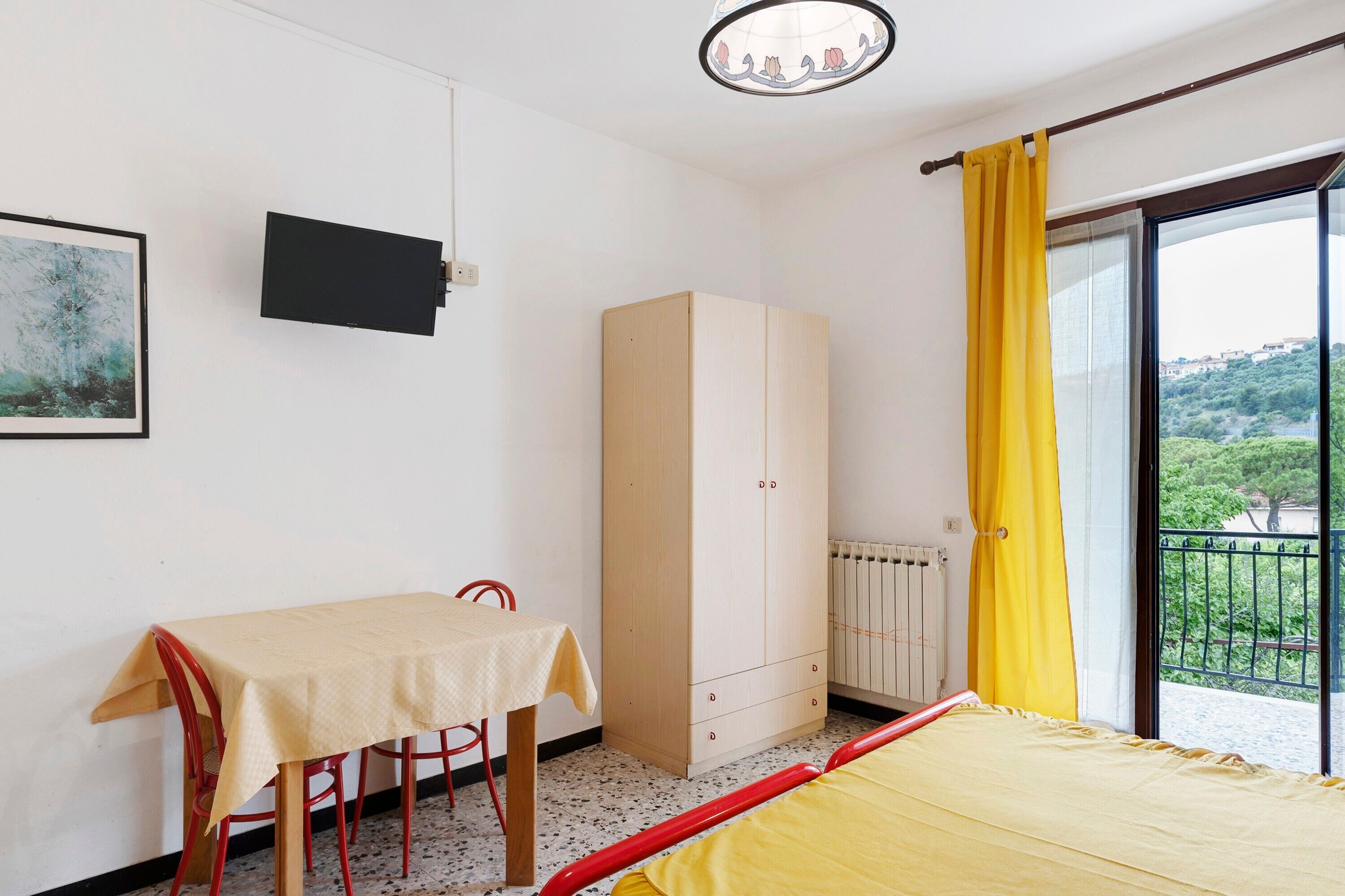 A bedroom with a bed, a table with chairs, a wardrobe, a radiator, and a window with a view of greenery.