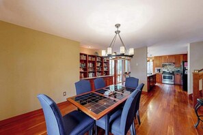 A dining area with a glass-top table, blue chairs, a chandelier, a bookshelf, and a kitchen in the background. - Spacious home near the ferry & downtown with enclosed yard, games, & W/D (Tisbury)