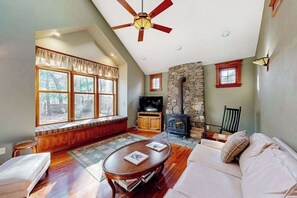 A cozy living room with a stone fireplace, a sofa, a television, and a large window with a view of trees. - Spacious home near the ferry & downtown with enclosed yard, games, & W/D (Tisbury)