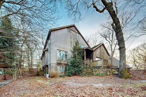 A rustic wooden house with a stone foundation, large windows, and a prominent tree in the foreground. - Spacious home near the ferry & downtown with enclosed yard, games, & W/D (Tisbury)