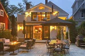 A two-story house with a patio area featuring outdoor furniture and a fire pit. - Luxury Gallery District home steps from beach with chef’s kitchen & gardens (Provincetown)
