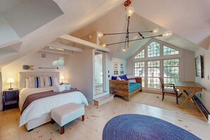 A bedroom with a bed, bedside table, lamp, and a view through large windows. - Luxury Gallery District home steps from beach with chef’s kitchen & gardens (Provincetown)