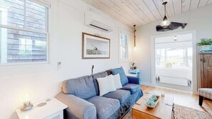 A room with a blue sofa, a white table, a wooden floor, a ceiling fan, and a window with blinds. - Renovated Waterfront Retreat | Inspiring Views | Walk to Beach, Dining, Shops (Provincetown)
