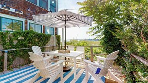 An outdoor patio with a table and chairs, an umbrella, and a view of a building and greenery. - Renovated Waterfront Retreat | Inspiring Views | Walk to Beach, Dining, Shops (Provincetown)