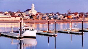 A waterfront marina with docked boats, a prominent church with a steeple, and residential buildings along the shore. - Renovated Waterfront Retreat | Inspiring Views | Walk to Beach, Dining, Shops (Provincetown)
