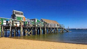 Un muelle con edificios que se extienden sobre el agua, sostenido por pilotes de madera, bajo un cielo azul despejado.