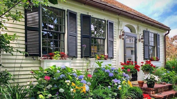 Un edificio blanco con contraventanas negras, un porche y un jardín con diversas flores.