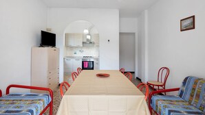 Zona pranzo con tavolo, sedie, televisore, armadio e cucina sullo sfondo.