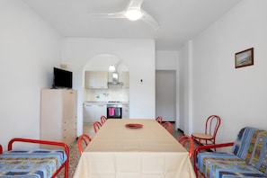 Un espace repas comprenant une table, des chaises, un téléviseur, une armoire et, au fond, une cuisine.