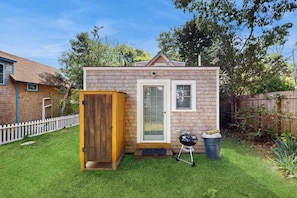 A small brick house with a wooden door, a window, and a barbecue grill in the yard.