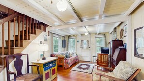 A spacious living area with a wooden staircase, a sofa, a chair, a small table, and a door leading to another room. - Cozy island cottage near local attractions with breezy yard & inviting interior (Oak Bluffs)