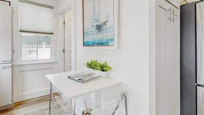 Een kamer met een witte tafel, een raam met zonwering, een lijst met een abstract schilderij, een potplant en een zwarte deur.