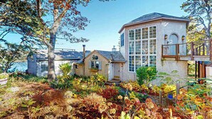 Una casa costera con un amplio ventanal, una pequeña construcción adosada y un jardín con diversas plantas.
