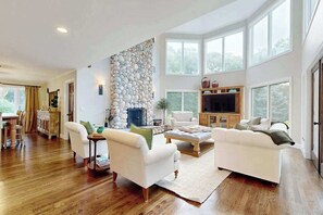 A spacious living room with a stone wall, a fireplace, a television, and large windows. - Enchanting home with tennis, firepit, golf nearby & washer/dryer (Oak Bluffs)