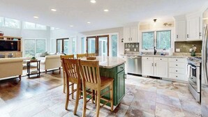 A spacious kitchen with a dining area, large windows, and a TV in the living room. - Enchanting home with tennis, firepit, golf nearby & washer/dryer (Oak Bluffs)