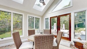 A sunroom with a large table and wicker chairs, large windows, and a view of the outdoors. - Enchanting home with tennis, firepit, golf nearby & washer/dryer (Oak Bluffs)