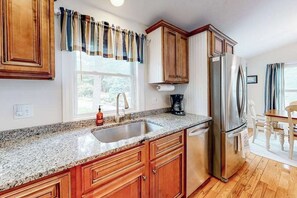 A kitchen with wooden cabinets, a stainless steel refrigerator, a granite countertop, and a window with curtains. - Cape escape with patio, grill, firepit, outdoor dining, W/D (Osterville)