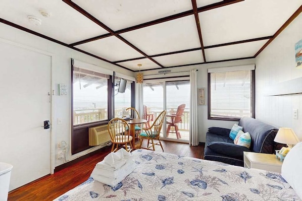 A room with a bed, a sofa, a dining area with chairs, and a view through large windows.