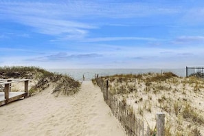 A sandy beach path leading to the ocean, with a wooden deck on the left and a fence on the right.