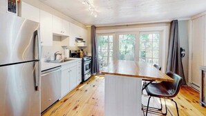 Una cocina moderna con armarios blancos, un refrigerador de acero inoxidable, una mesa de comedor de madera con una silla y un amplio ventanal con cortinas.