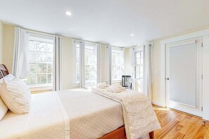 A bedroom with a large bed, multiple windows, and a door with a shutter. - Historic, dog-friendly beach view house with fast WiFi, ocean view, & grill (Oak Bluffs)