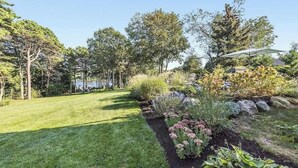 Ein gepflegter Garten mit verschiedenen Pflanzen und Bäumen, ein klarer Himmel und ein Haus im Hintergrund.