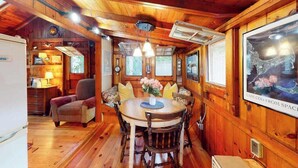A wooden cabin interior with a dining table, chairs, a sofa, a lamp, and a painting on the wall. - Drummer Cove Gem | Cottage in Wellfleet with patio, steps from the cove (Wellfleet)