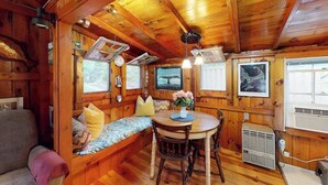 A cabin interior with a wooden bench, a dining table with chairs, a clock, and a window with a view. - Drummer Cove Gem | Cottage in Wellfleet with patio, steps from the cove (Wellfleet)