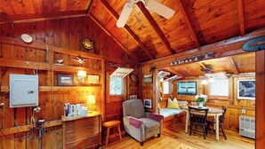 A wooden cabin interior with a dining area, a chair, a small table, and a ceiling fan. - Drummer Cove Gem | Cottage in Wellfleet with patio, steps from the cove (Wellfleet)