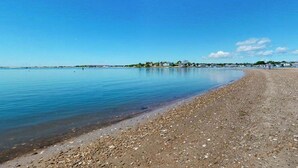 A beach with calm water, a pebbly shore, and a distant shoreline with buildings and greenery. - Coastal bay view home with patio, W/D, AC, & amazing location (West Yarmouth)