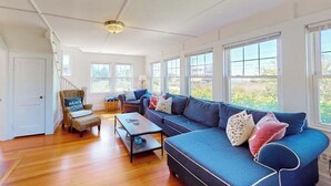 A spacious living room with a large blue sofa, a wooden coffee table, and a wicker chair with a pillow labeled 'BEACH HOUSE'. - Coastal bay view home with patio, W/D, AC, & amazing location (West Yarmouth)