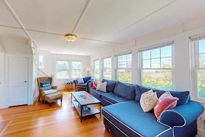 A spacious living room with a large blue sofa, a wooden coffee table, and a wicker chair with a pillow labeled 'BEACH HOUSE'. - Coastal bay view home with patio, W/D, AC, & amazing location (West Yarmouth)