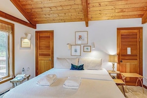 A room with a wooden ceiling, a bed with white linens, a bedside table with a lamp, and framed pictures on the wall. - Charming 2-story house near the beach with inviting deck, large yard & canoe (West Tisbury)