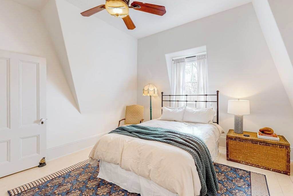 Un dormitorio con cama, ventilador de techo, ventana, mesita de noche con lámpara y una alfombra decorativa.