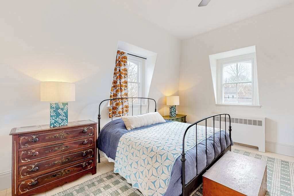 A bedroom with a bed, dresser, two bedside lamps, a window, and a radiator.