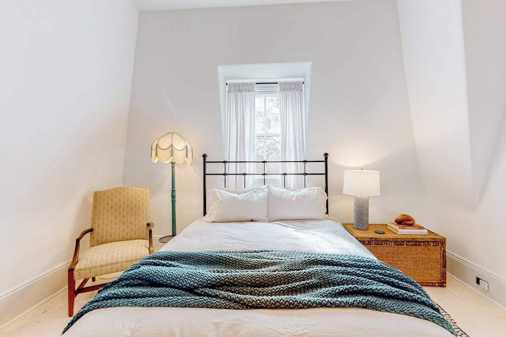 Un dormitori amb un llit, una cadira, una taula de nit amb una làmpada i una finestra amb cortines.