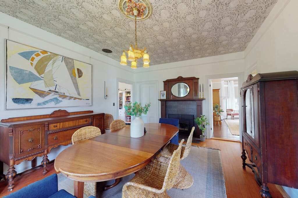 Uma sala de jantar com mesa de madeira, cadeiras de vime, lareira e uma grande pintura de um veleiro na parede.