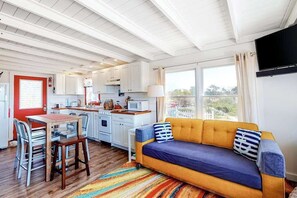 Eine kompakte Küche mit weißen Schränken, ein Essbereich mit Tisch und Stühlen, ein Sofa mit Kissen und ein an der Wand montierter Fernseher.