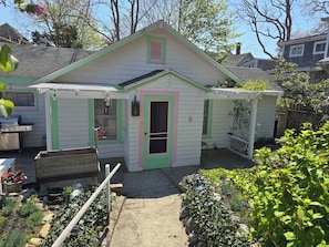 A small, single-story house with a green door and pink trim, a covered patio, and a garden with various plants.