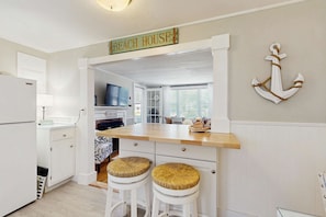 Une cuisine avec un plan de travail en bois, des meubles blancs, deux tabourets en osier, une enseigne indiquant « MAISON DE PLAGE » et une ancre décorative accrochée au mur.