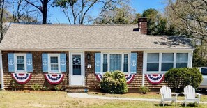 A house with a brick exterior, white trim, and blue shutters. There are two white chairs outside. - Cozy Cape Cottage w/ large yard, outdoor shower & close to beaches & attractions (West Yarmouth)