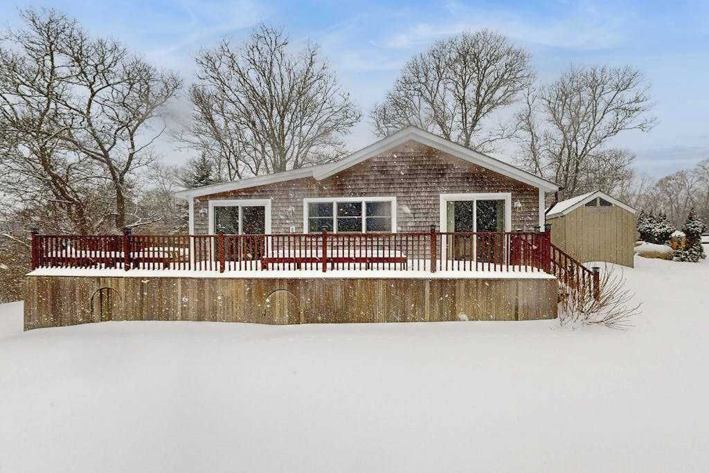 A wooden cabin with a fenced deck, surrounded by snow-covered ground and trees.