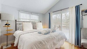 Un dormitorio con cama, mesita de noche, lámpara, ventana con persianas y vistas al exterior.