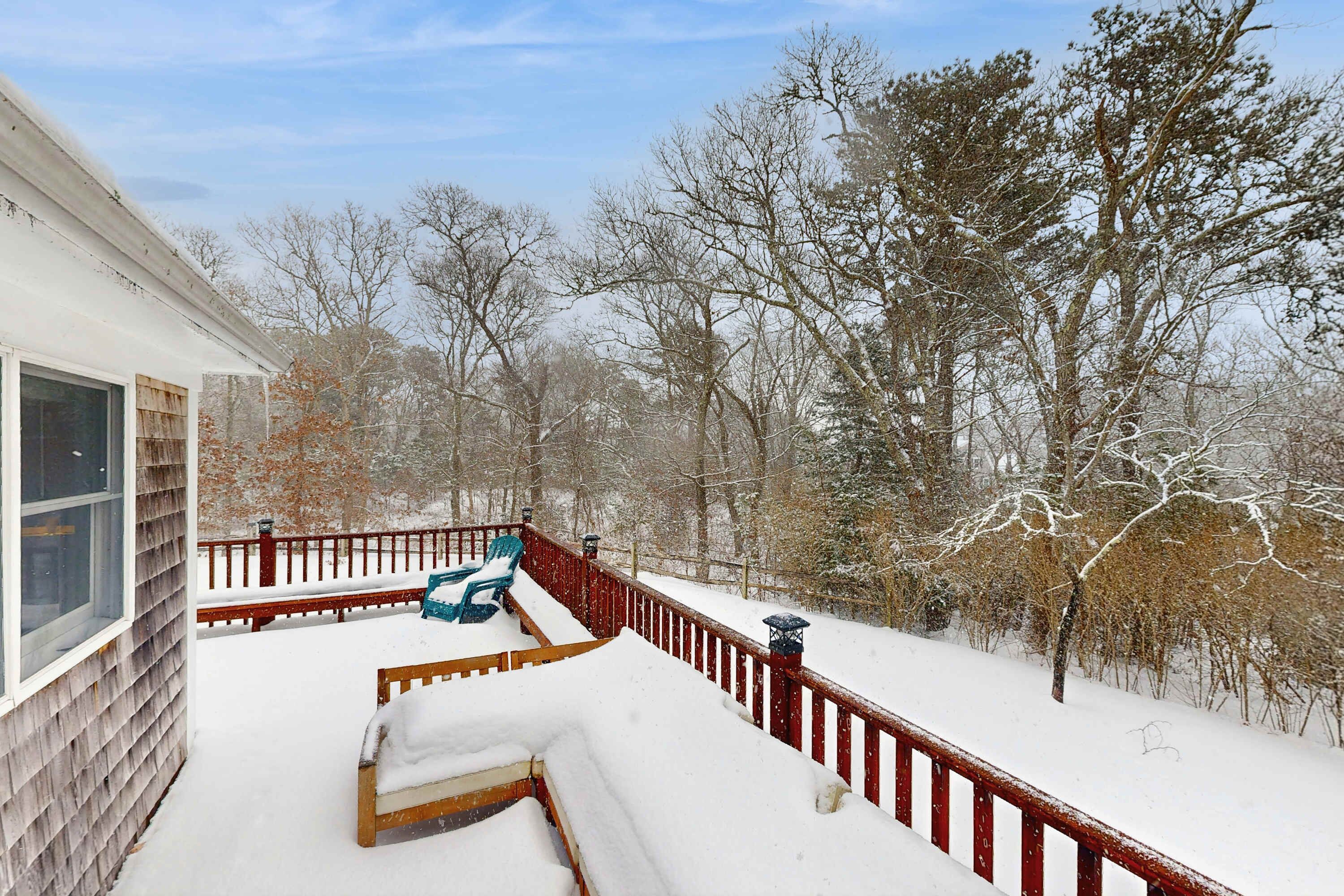 Um deck de madeira com móveis cobertos de neve e um corrimão, rodeado por árvores.