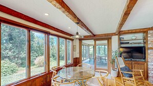 A room with large windows, a round glass table, wicker chairs, a wooden ceiling, and a fireplace.