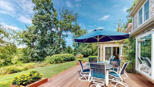 A deck with outdoor furniture and a large umbrella, surrounded by greenery and a house.