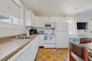 A compact kitchen with white cabinets, a microwave, a stove, a refrigerator, a sink, and a dining area with a table and chairs. - Downtown studio w/ full kitchen - walk to Bay and shops! (Provincetown)