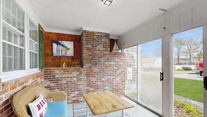 Ein Zimmer mit Backsteinwand, einem Korbssessel mit Kissen, einem Holztisch und einer Glastür zum Außenbereich.