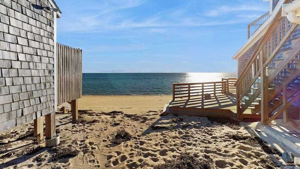 Ein Strand mit einer Holzterrasse, die zum Meer führt, ein Gebäude mit Satteldach und ein klarer Himmel.