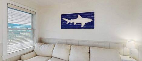 Ein Zimmer mit einem weißen Sofa, einer Wanddekoration in Form eines Hai-Silhouettenbildes, einem Fenster mit Jalousien und einem Nachttisch mit Lampe.